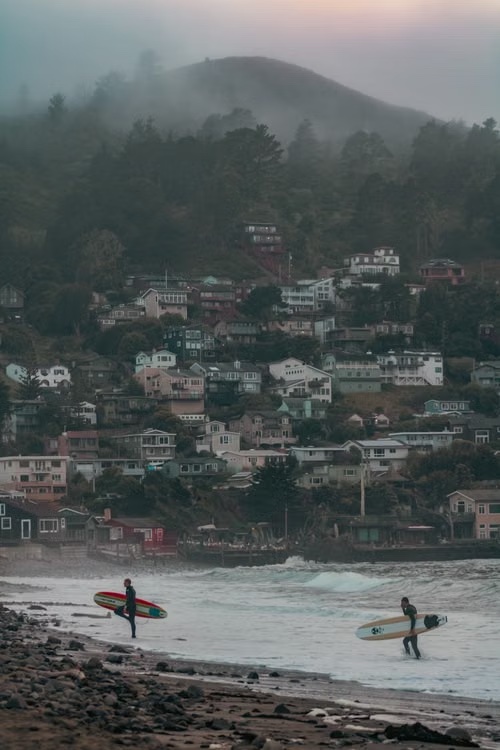 Surfers walking out of the water below a foggy hillside town on the Northern California coast